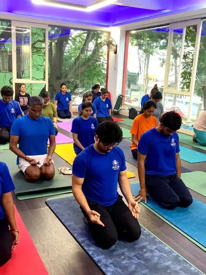 A group of students in matching shirts sit in Vajrasana, ready for a meditation or pranayama session.