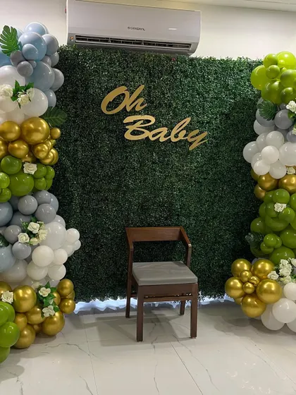 An elegant "Oh Baby" shower setup with a lush green foliage wall. The backdrop is framed by beautiful balloon columns in shades of green, gold, grey, and white, creating a fresh and natural look.