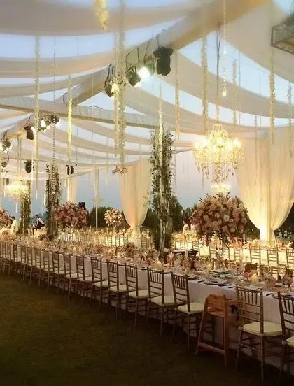 An exquisite long-table dining setup for an outdoor wedding reception. The tables are arranged under a canopy of white drapes and hanging floral strings, with chandeliers adding a touch of elegance for an evening under the stars.