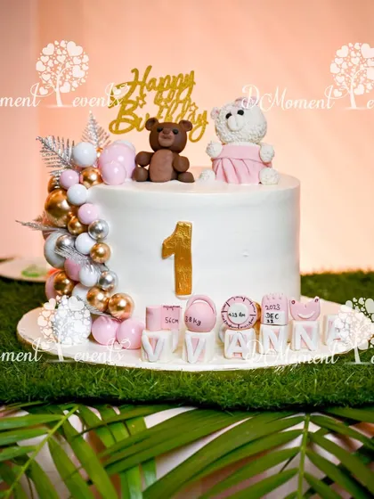 An elegant first birthday cake for a teddy bear theme, featuring two cute teddy bears and the baby's name spelled out in decorative blocks.