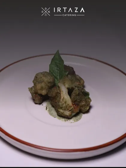 My signature herb-crusted cauliflower starter. The dish is delicately plated with a smear of green chutney and a fresh basil leaf, highlighting the fresh, earthy flavors.