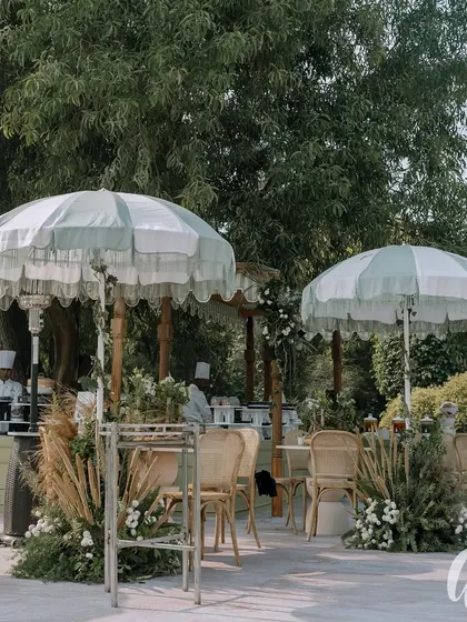 An outdoor seating area designed for a classic ivory and green themed wedding. Fringed umbrellas and comfortable seating are surrounded by arrangements of pampas grass and white florals.