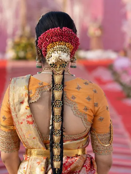 A stunning view of a bride's back, showcasing the scalloped cutout of her orange blouse and elaborate hair adornments. The blouse embroidery perfectly matches the traditional aesthetic of her wedding look.