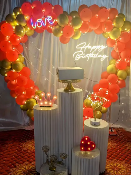 A romantic heart-shaped balloon arch in red and gold for a birthday celebration. The setup includes a 'Love' neon sign and other light-up props.
