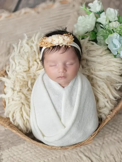 A newborn girl wrapped in white, resting in a rustic basket with soft flowers.