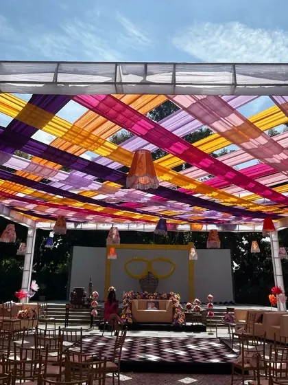 A large-scale carnival setup with a ceiling of woven multi-colored ribbons, creating a playful and festive atmosphere for a daytime event.