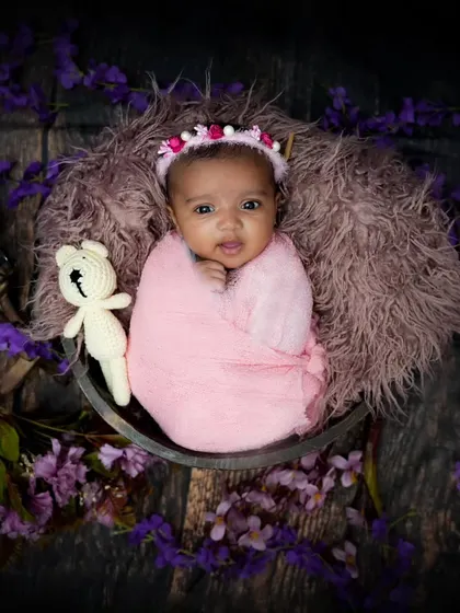 An awake and alert newborn wrapped in pink, surrounded by purple flowers. Her direct gaze is captivating.