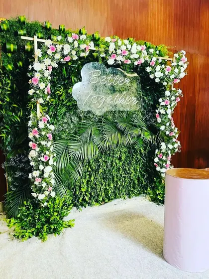 This greenery wall photo booth is framed with an arch of pink and white flowers. A custom "Better Together" neon sign adds a modern and personal touch, perfect for a wedding anniversary or engagement party.