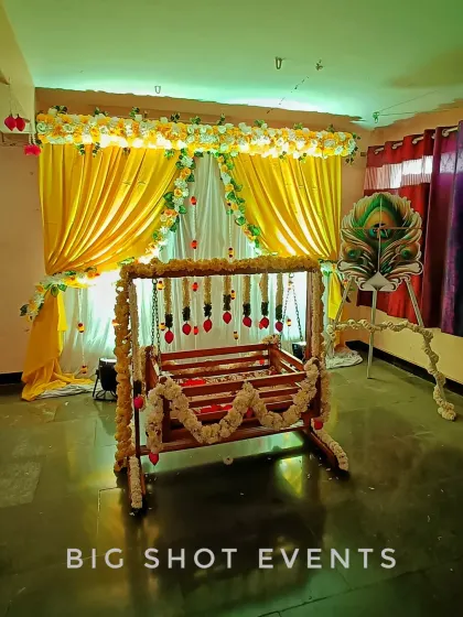 A traditional cradle ceremony with a touch of yellow and white. The backdrop features bright yellow drapes, and the wooden cradle is decorated with delicate flower strings. A peacock feather prop adds a symbolic and auspicious element to the decor.