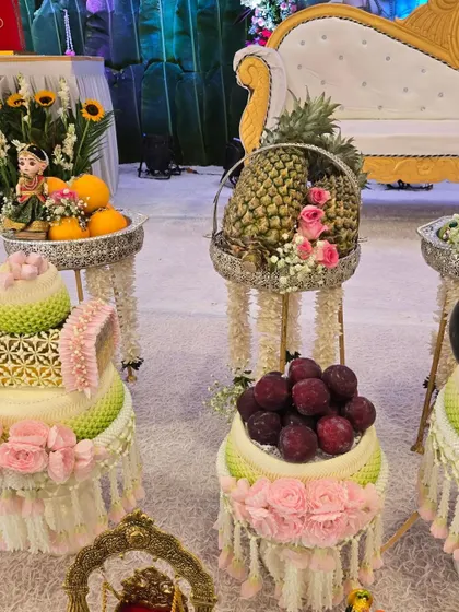 An elegant display of fresh fruits and traditional Thai floral arrangements on decorative stands for a baby shower.
