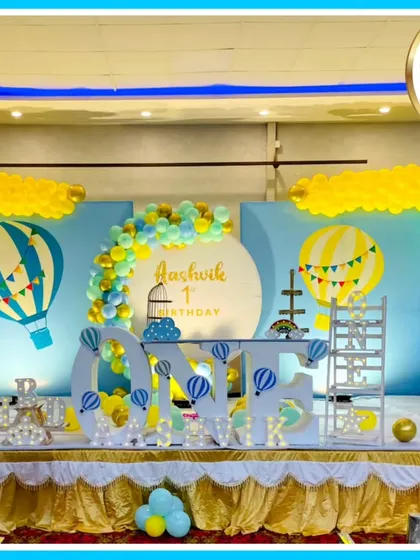 A breathtaking hot air balloon theme for a first birthday. This expansive stage setup includes a large 'ONE' prop, multiple hot air balloon models, cloud and sky backdrops, and extensive balloon work in a cheerful blue and yellow palette.