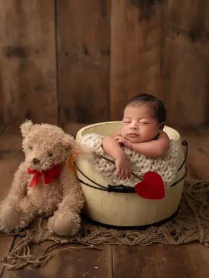 A sweet newborn boy in a bucket, accompanied by a classic teddy bear. The rustic wood background adds warmth to this adorable portrait.