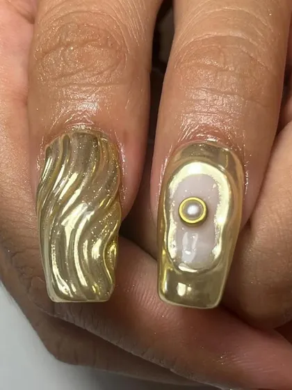 A macro shot of our textured gold chrome nails. One nail features a 3D wave pattern, while the other has a single pearl set in a gold bezel, like a piece of jewelry.
