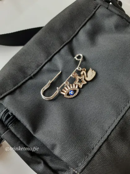 Another custom safety pin charm, this time with an evil eye, a star, and a swan, attached to a black crossbody bag. It's a great way to personalize your accessories.