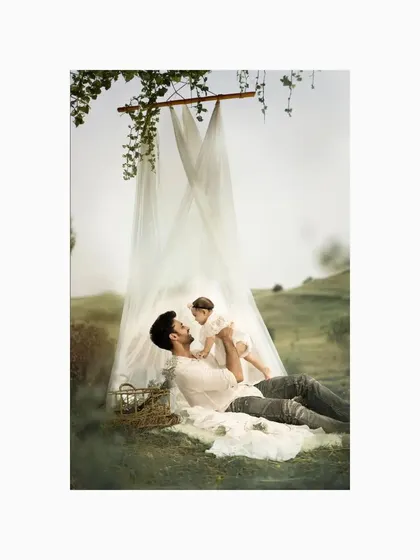 In a whimsical outdoor setting, a father lifts his baby girl under a canopy of sheer fabric and vines. This creative baby portrait captures the unbreakable bond and playful love between a father and his daughter.