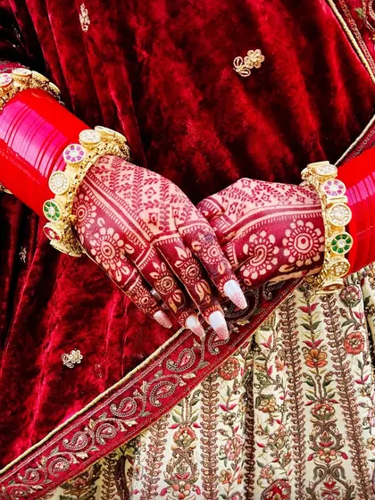A close up of a bride's hands, showing the beautiful floral mehndi stain against her red and cream lehenga. The color is a perfect, classic bridal red.