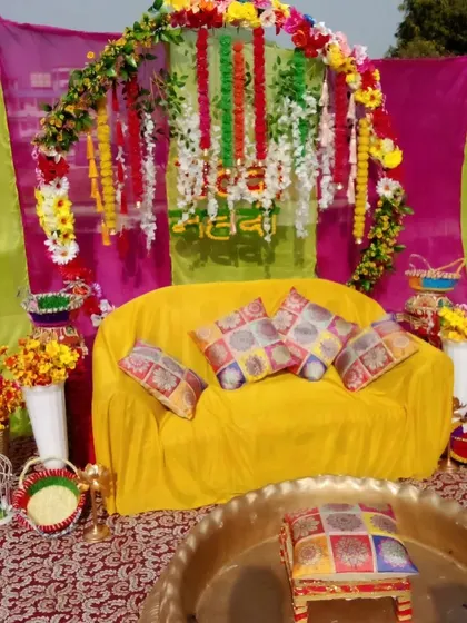 An outdoor Mehendi setup with a simple floral arch over a yellow sofa. The decor uses a mix of colorful drapes and props like birdcages for a rustic feel.