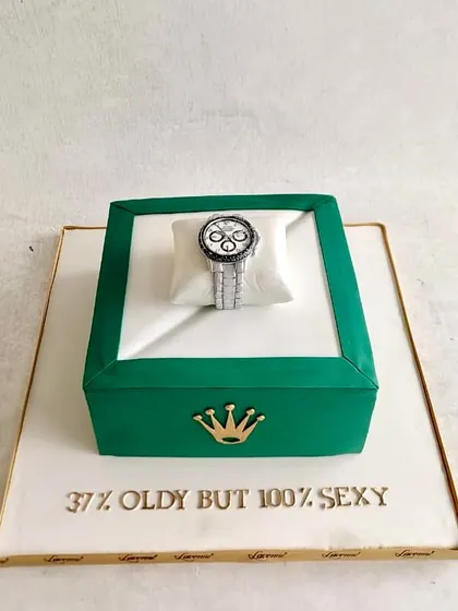 A luxurious Rolex-themed cake. The green watch box is the cake, and it's topped with a detailed, edible Rolex watch resting on a sugar pillow. A perfect cake for a milestone birthday.