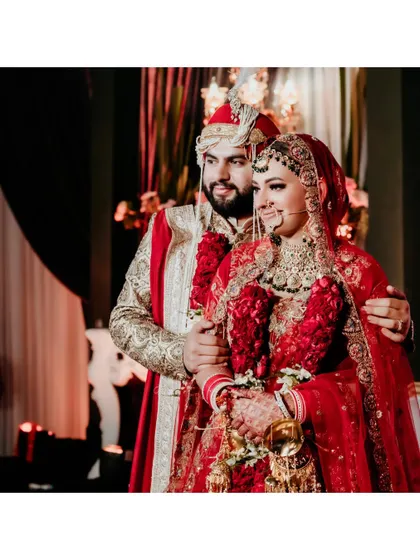 A classic portrait of the couple in their wedding attire. The bride's red lehenga and the groom's gold and red sherwani are perfectly coordinated.