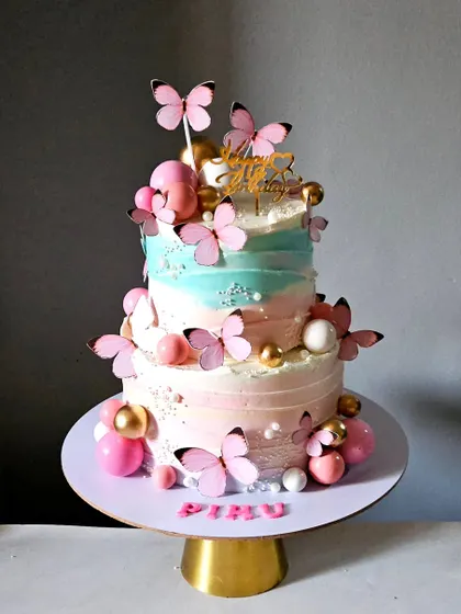 A three-tiered cake featuring a beautiful gradient of pink and blue whipped cream. The design is adorned with pink butterflies and spheres in shades of pink and gold, creating a graceful and sweet birthday cake.