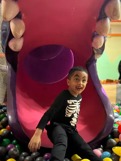 A boy in a skeleton costume flashes an excited smile after coming down the big purple slide into the ball pit.