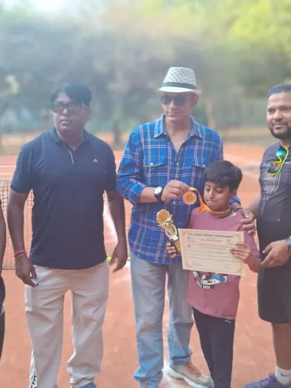 A young player receiving his medal and certificate for the U12 Aptitude Title at the Ideal Tennis Open Tournament, surrounded by coaches and officials on the clay court.