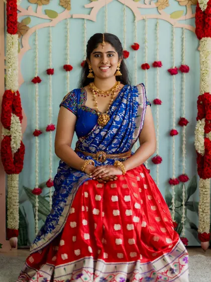 A bride in a vibrant half-saree for a traditional ceremony. The backdrop features intricate floral work that complements her attire perfectly.
