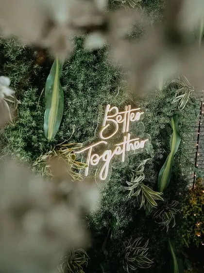 Another perspective of the "Better Together" neon sign, seen through a foreground of white flowers, adding to the enchanted forest feel.