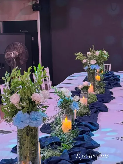 This wider shot of the long dining table highlights the full tablescape design. The combination of fresh greenery, blue and white flowers, and glowing candles creates a serene and elegant setting for a special dinner.