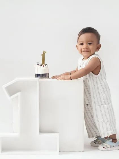 A little boy proudly stands next to a large '1' prop and his first birthday cake in a clean, minimalist all-white studio setting.