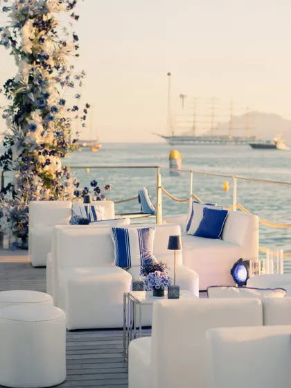 The lounge setup for the welcome dinner, with comfortable white sofas, blue and white striped cushions, and a floral arch, all set against the golden hour glow on the water.