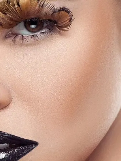 A macro shot of the model's skin and eye, showing the intricate detail of the feather makeup. The lighting reveals natural skin texture while maintaining a polished, high-end feel.
