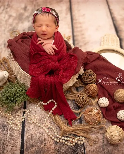 A rich and rustic setup featuring a baby in a deep red wrap. The wooden background, pearls, and wicker props create a beautifully textured and warm portrait.