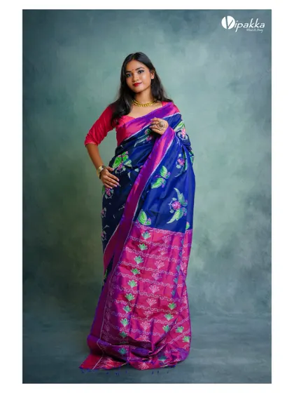 A studio shot for a saree brand. The model's pose displays the full length and vibrant colors of the blue and pink silk saree against a textured studio background.