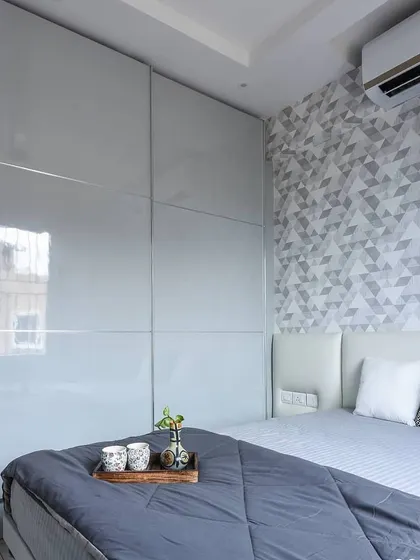 A contemporary bedroom featuring a geometric-patterned wallpaper accent wall. The glossy light grey sliding wardrobe provides a sleek and unobtrusive storage solution.
