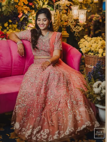 A smiling bride seated on a vibrant pink sofa, her elaborate peach lehenga spread out beautifully.
