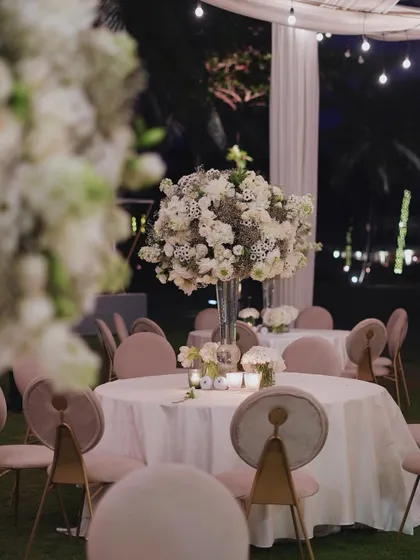 As evening falls, the same reception setup is transformed by soft lighting. The large white floral centerpieces seem to glow in the twilight, creating a romantic and intimate atmosphere for the dinner celebration.