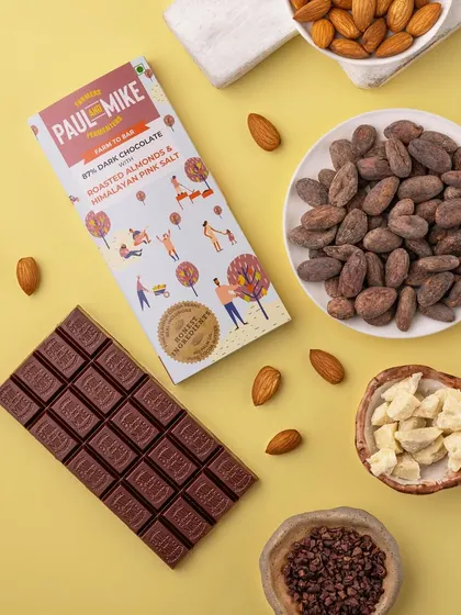 A bright and clean shot of an almond chocolate bar on a yellow background, with raw ingredients like cocoa beans and almonds.
