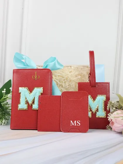 A close-up of the red monogrammed travel set, perfect for Rakhi. The set includes a passport cover, card holder, and luggage tag, all personalized with a chenille patch initial.