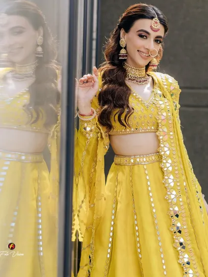 A joyful bride in a vibrant yellow mirror-work lehenga, her smile reflected in the glass beside her, creating a bright and happy portrait.