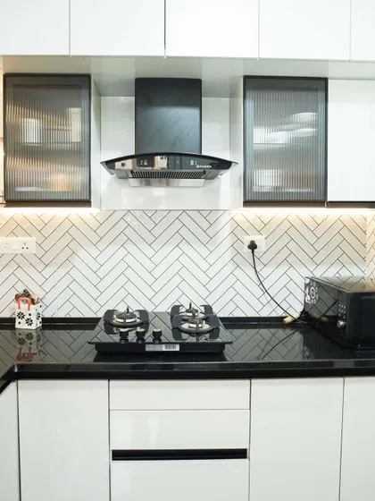 A minimalist kitchen design featuring all-white handleless cabinets and a striking herringbone tile backsplash. The black granite countertop and integrated chimney add a touch of modern contrast.