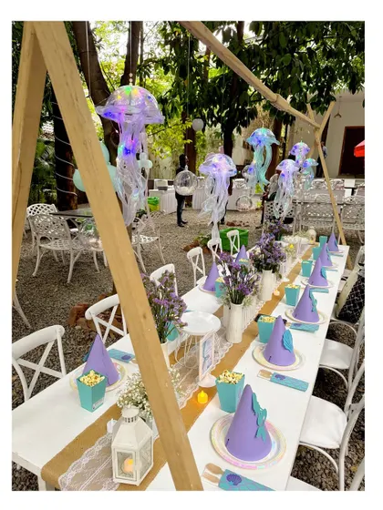 A stunning kids' dining table setup for a mermaid party. The table is decorated with hanging jellyfish, themed party hats, and a lace runner, creating an elegant underwater feast.