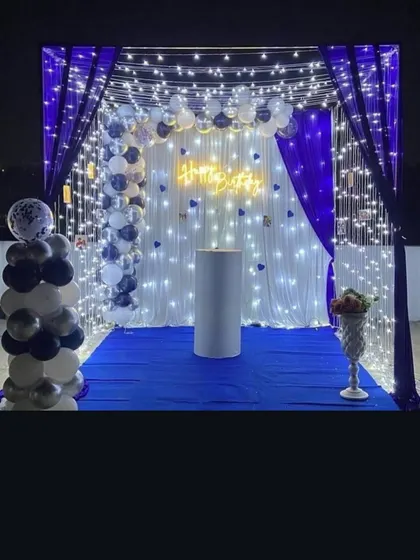 A stunning canopy-style birthday decoration for an outdoor or terrace party. The setup uses white and blue drapes with fairy lights, a neon sign, and balloon pillars.