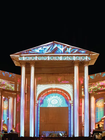 The monumental stage is transformed by vibrant projection mapping, turning the classical architecture into a canvas of light and color.