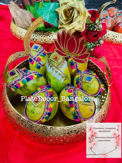 Green mangoes decorated with vibrant, multi-colored lace and kundan stones in a golden basket. The lotus cutout adds a traditional touch.