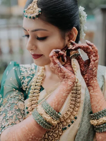A close-up of a bride getting ready, wearing an off-white saree with a beautifully embroidered green blouse. The sheer puff sleeves are a delicate touch.