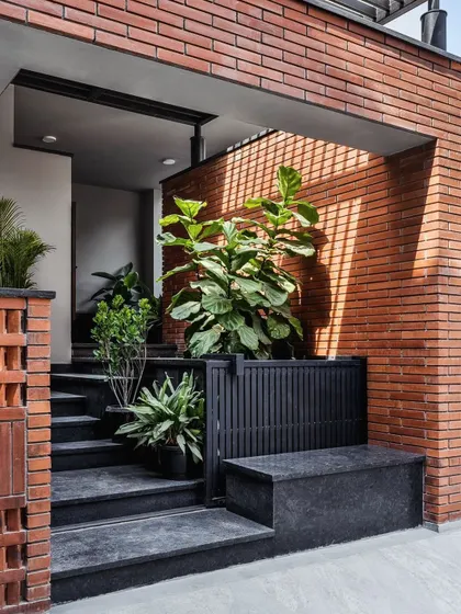 The entrance to this home features a crisp combination of red brick, black granite, and lush plants. The play of light through the brick jaali wall creates a welcoming pattern of shadows, guiding you into the space.