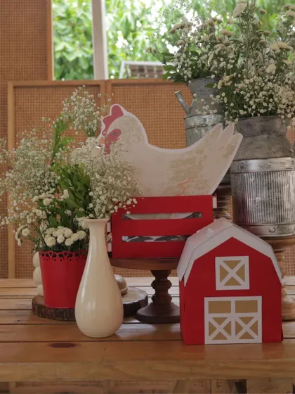A close-up of a farm-themed centerpiece, featuring a hen cutout in a red crate, a miniature barn, and fresh baby's breath in milk cans and vases.