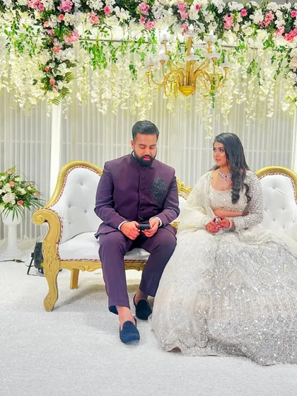 A candid moment on a beautifully decorated stage. The couple is seated on an elegant white sofa, with a backdrop of hanging white wisteria and a lush floral border.
