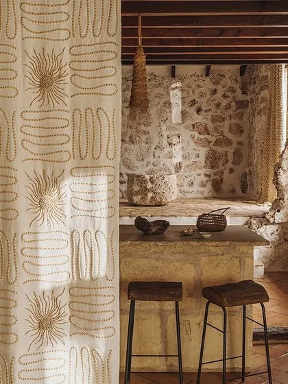 The 'Perpétuel Soleil' fabric used as a curtain in a rustic, stone-walled interior. The embroidered design adds a layer of artisanal charm, perfectly complementing the natural textures of the space.